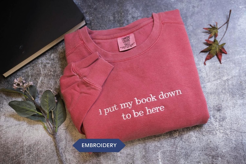 a red long-sleeved shirt with the text "I put my book down to be here" printed on it, along with a black book and some leaves.