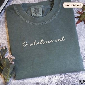 May include: A blue spruce t-shirt with the embroidered phrase "to whatever end" in white script. The shirt is folded and has a Comfort Colors tag. The image includes decorative leaves and the word "Embroidered".