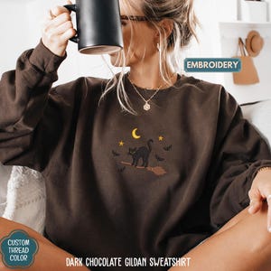 A woman wearing a brown sweater with a cat embroidered on it, holding a mug, and sitting on a couch.