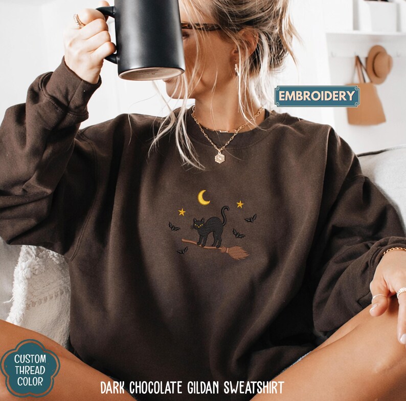 A woman wearing a brown sweater with a cat embroidered on it, holding a mug, and sitting on a couch.