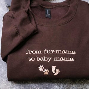 A brown sweatshirt with the text "from fur mama to baby mama" and paw prints is displayed on a gray surface.