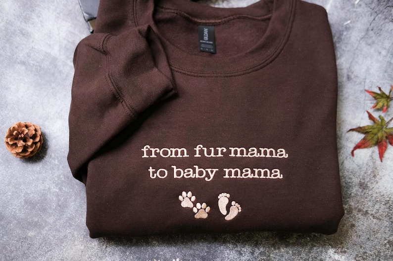 A brown sweatshirt with the text "from fur mama to baby mama" and paw prints is displayed on a gray surface.