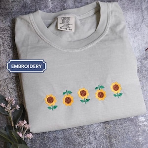 a gray t-shirt with a row of embroidered sunflowers on the front, placed on a gray surface alongside some leaves and flowers.