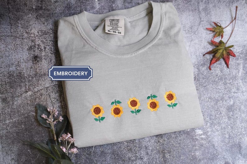 a gray t-shirt with a row of embroidered sunflowers on the front, placed on a gray surface alongside some leaves and flowers.