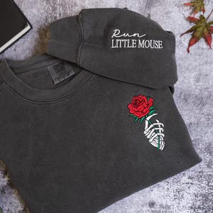 a gray sweatshirt with a rose embroidered on the front, a gray cap with the text "Run Little Mouse" on it, and some leaves are visible in the background.