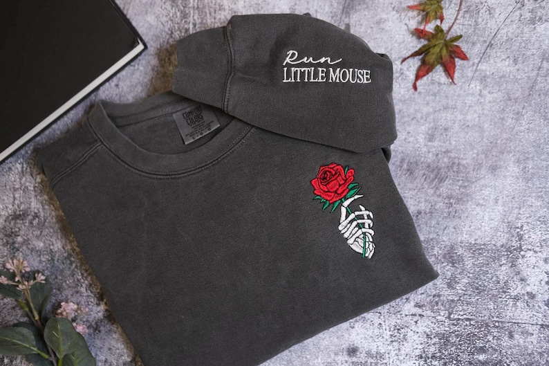 a gray sweatshirt with a rose embroidered on the front, a gray cap with the text "Run Little Mouse" on it, and some leaves are visible in the background.