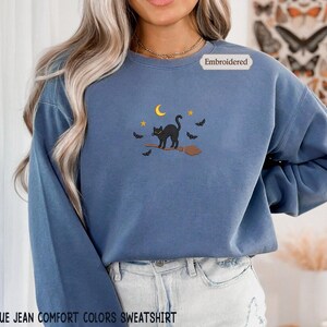 A woman wearing a blue sweatshirt with a black cat embroidered on it, standing in front of a floral background.