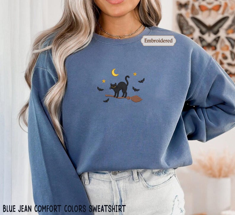 A woman wearing a blue sweatshirt with a black cat embroidered on it, standing in front of a floral background.