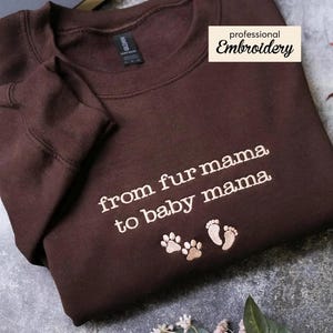 A brown sweatshirt with the text "from fur mama to baby mama" and paw prints, along with a label that says "professional Embroidery" and a pine cone.