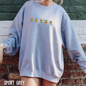 A person wearing a grey sweatshirt with a row of yellow sunflowers printed on it, standing in front of a brick wall.