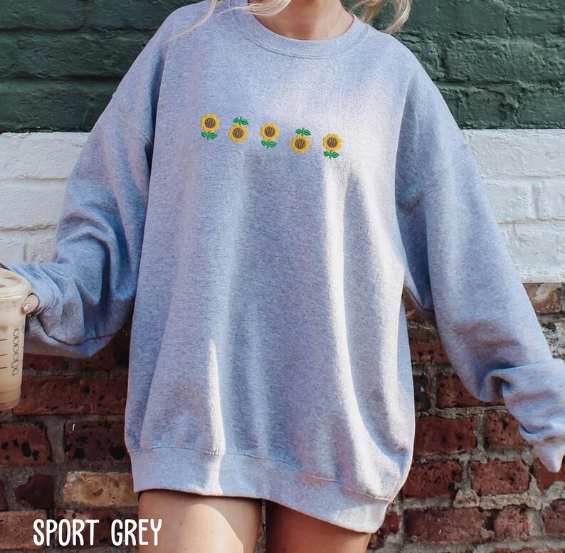 A person wearing a grey sweatshirt with a row of yellow sunflowers printed on it, standing in front of a brick wall.