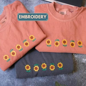 two pink t-shirts with yellow sunflowers embroidered on them, laid out on a gray surface.