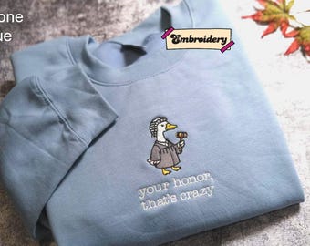 May include: A stone blue crewneck sweatshirt featuring an embroidered graphic of a duck dressed as a judge, holding a wooden gavel. Below the duck, the text reads, "your honor, that's crazy" in white thread. The garment has long sleeves.