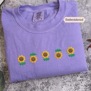 A purple t-shirt with a row of embroidered sunflowers on it, along with a label indicating that it is embroidered.