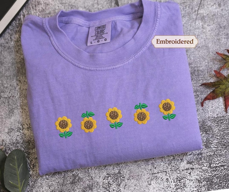A purple t-shirt with a row of embroidered sunflowers on it, along with a label indicating that it is embroidered.