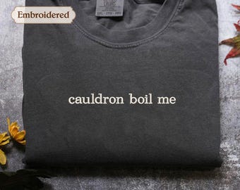 May include: A charcoal gray crew neck t-shirt featuring the embroidered text "cauldron boil me" in white thread across the chest. The shirt has a Comfort Colors brand label at the inner neckline indicating a size 2XL.