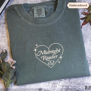 May include: A blue spruce t-shirt with the word "Embroidered" in the upper right corner. The shirt features a white embroidered heart design with the words "Midnight Reader" and a book illustration. The shirt is folded and has a Comfort Colors tag.