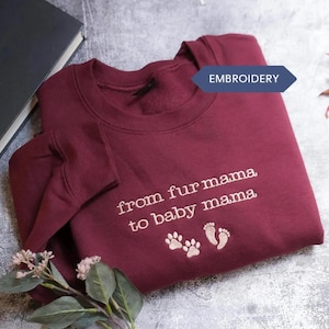 A maroon sweatshirt with the text "from fur mama to baby mama" and paw prints is displayed on a gray surface, accompanied by a black book and a small plant.