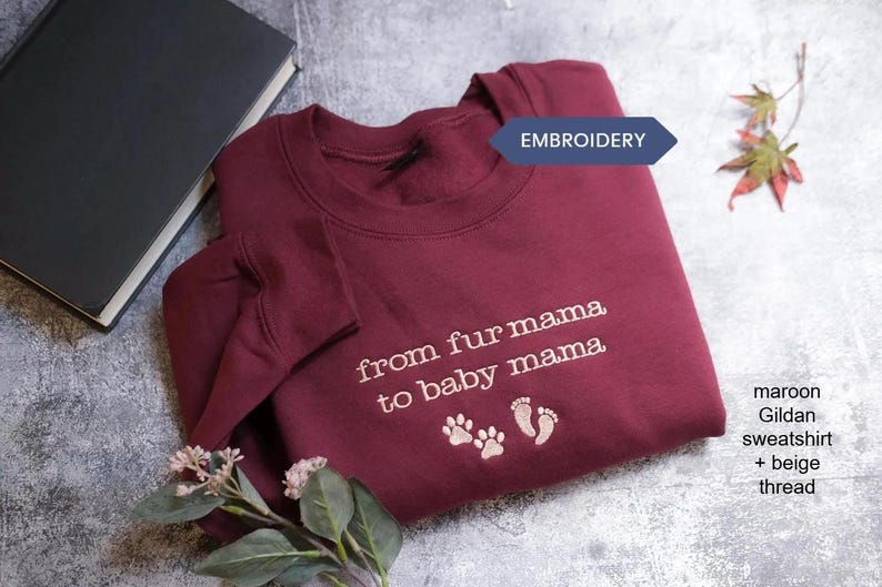 A maroon sweatshirt with the text "from fur mama to baby mama" and paw prints is displayed on a gray surface, accompanied by a black book and a small plant.