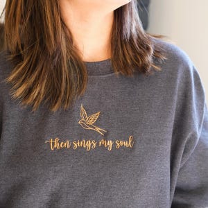 May include: A white ceramic mug with a black handle and the text "But First, Coffee" in a bold, black font. The mug is set against a blurred background, suggesting a cozy setting.