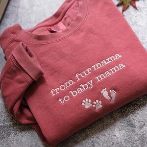 A red sweatshirt with the text "from fur mama to baby mama" and paw prints is laid out on a gray surface.
