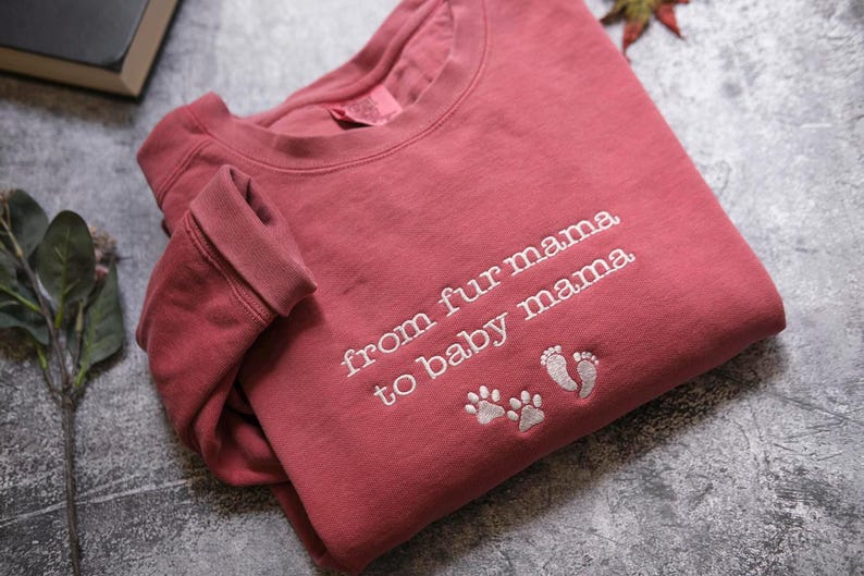 A red sweatshirt with the text "from fur mama to baby mama" and paw prints is laid out on a gray surface.