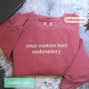 May include: A dusky pink sweatshirt with the text "your custom text embroidery" in white. The sweatshirt has a crew neck and long sleeves. The tag reads "Comfort Colors" and the word "Embroidered" is in a tag on the top right.
