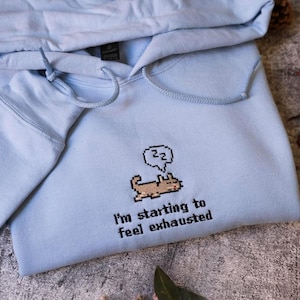 May include: A light blue hoodie with a drawstring hood. The front features an embroidered pixelated dog with a speech bubble and the text "I'm starting to feel exhausted."