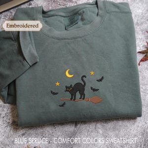 A gray sweatshirt with a black cat embroidered on it, along with a crescent moon, stars, and bats, is displayed on a gray surface.