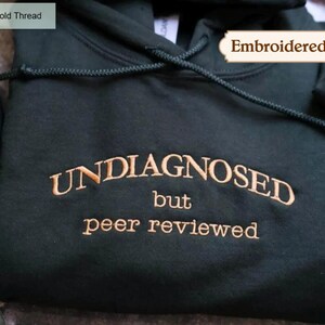 May include: Dark green hoodie with gold thread embroidery. The text "UNDIAGNOSED but peer reviewed" is embroidered across the chest. The hoodie features a drawstring hood and is made of a soft fabric.