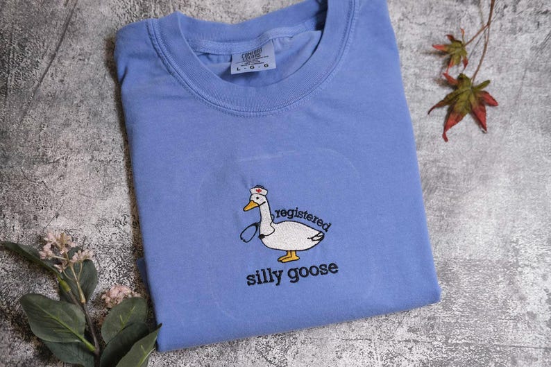 a blue t-shirt with a cartoon goose and the text "registered silly goose" printed on it, along with some leaves and flowers on a textured surface.