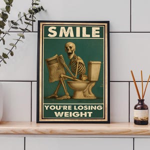May include: Framed poster with a green background featuring a skeleton sitting on a toilet, reading a newspaper. The poster has the words "SMILE" at the top and "YOU'RE LOSING WEIGHT" at the bottom, in white text.