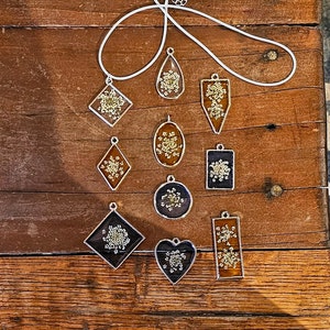 Könnte beinhalten: Eine Sammlung von 10 silbernen Halsketten mit verschiedenen Anhängerformen. Jeder Anhänger zeigt ein gepresstes Blumenmotiv mit einer klaren Harzbeschichtung. Die Anhänger haben verschiedene Formen, darunter quadratisch, oval, tropfenförmig, herzförmig und rechteckig.