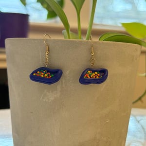 Pode incluir: Um par de brincos feitos à mão com um design em forma de tigela azul escuro preenchido com granulado redondo e colorido. Os brincos têm ganchos dourados e são exibidos em um fundo neutro.