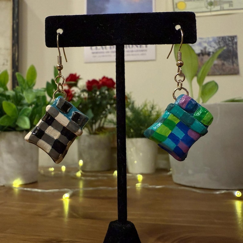 May include: Handmade earrings with a woven basket design. One earring has a black, white, and grey chequered pattern, the other a vibrant mix of green, blue, and purple squares. Both hang from gold-coloured hooks.
