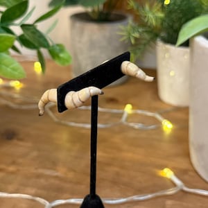 May include: A pair of croissant-shaped earrings, displayed on a black stand. The earrings are a light beige colour with detailed lines. The background features greenery and fairy lights.