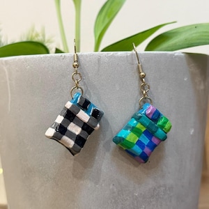 May include: A pair of handmade earrings with a woven basket design. One earring features a black, white, and gray checkered pattern, while the other showcases a vibrant mix of blue, green, and purple squares, suspended from gold-colored hooks.