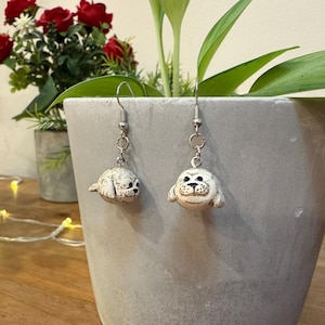 May include: A pair of seal-shaped earrings. Each earring features a small, white seal with black details, suspended from a silver-colored hook. The seals have a textured appearance and are detailed with eyes, noses, and whiskers. The earrings are displayed against a neutral background.