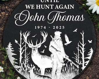 May include: A round black memorial stone with the text "UNTIL WE HUNT AGAIN" and the name "John Thomas" with the years 1974-2025. The design features a hunter, deer, and trees in a forest scene.