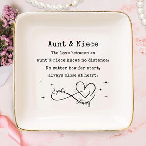 May include: A white ceramic trinket dish with gold trim. The dish features the text "Aunt & Niece" and a heartfelt message about the bond between an aunt and niece. Includes the names "Sophia" and "Jenny" with a heart and infinity symbol.