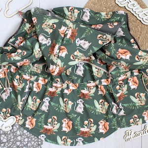 Könnte beinhalten: Grüne Kapuzenjacke mit Waldmuster, das Eichhörnchen, Pilze und Blätter zeigt. Die Jacke hat einen Kordelzug in der Taille, einen Reißverschluss und graue Bündchen. Thema: Kinderkleidung und Natur.