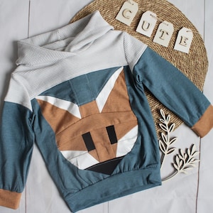 Könnte beinhalten: Blauer und brauner Kapuzenpullover mit einem Fuchskopf-Design auf der Vorderseite. Der Pullover hat eine weiß-grau gestreifte Kapuze und braune Bündchen. Das Wort "CUTE" steht auf Holzanhängern.