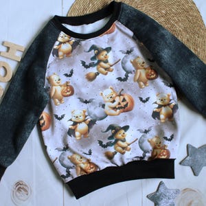 Könnte beinhalten: Ein langärmliges Halloween-Sweatshirt. Der Körper des Shirts zeigt ein Muster aus Cartoon-Bären in Halloween-Kostümen, Kürbissen und Fledermäusen. Die Ärmel sind dunkelgrau, die Bündchen und der Halsausschnitt sind schwarz.