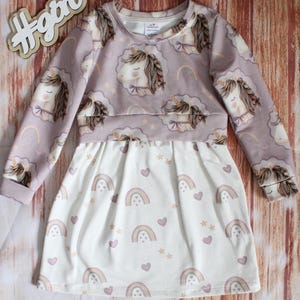 Könnte beinhalten: Ein langärmeliges Kleid mit einem malvenfarbenen Oberteil mit wiederkehrendem Einhorn- und Regenbogenmuster, kombiniert mit einem weißen Rock, der mit kleinen Regenbögen, Sternen und Herzen bedruckt ist. Das Kleid hat einen Rundhalsausschnitt und Rippbündchen.