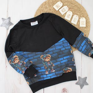 Könnte beinhalten: Schwarzes Langarm-Sweatshirt mit blauem Ziegelmuster und Teddybär-Grafiken. Die Ärmel und der untere vordere Teil zeigen das Bären-Design. Der obere Teil des Sweatshirts ist einfarbig schwarz.