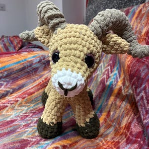 May include: A crocheted ram toy with a tan body, grey horns, and dark brown hooves. It has large black eyes and a white muzzle. The toy is placed on a colourful, patterned blanket.