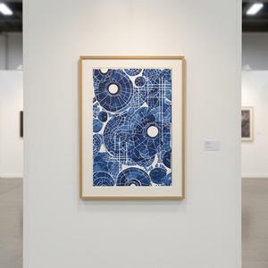 May include: A framed abstract art print featuring a pattern of blue and white overlapping circles and lines. The artwork is displayed on a white wall in a gallery setting, with a wooden frame.