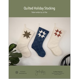 Puede incluir: Tres calcetines navideños acolchados en crema y azul marino cuelgan de una pared blanca. Cada calcetín presenta un diseño de estrella en marrón, blanco y rojo. El texto "Quilted Holiday Stocking" y "Pattern written by Juli Rae" es visible.