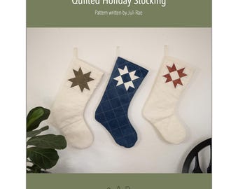 May include: Three quilted holiday stockings in cream and navy blue hang against a white wall. Each stocking features a star design in brown, white, and red. The text "Quilted Holiday Stocking" and "Pattern written by Juli Rae" are visible.