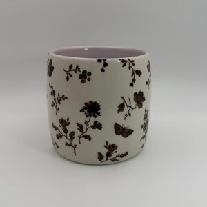 May include: A ceramic tumbler with a cream-colored exterior adorned with brown floral and butterfly patterns. The interior rim is a soft pink. The tumbler is cylindrical and suitable for beverages.